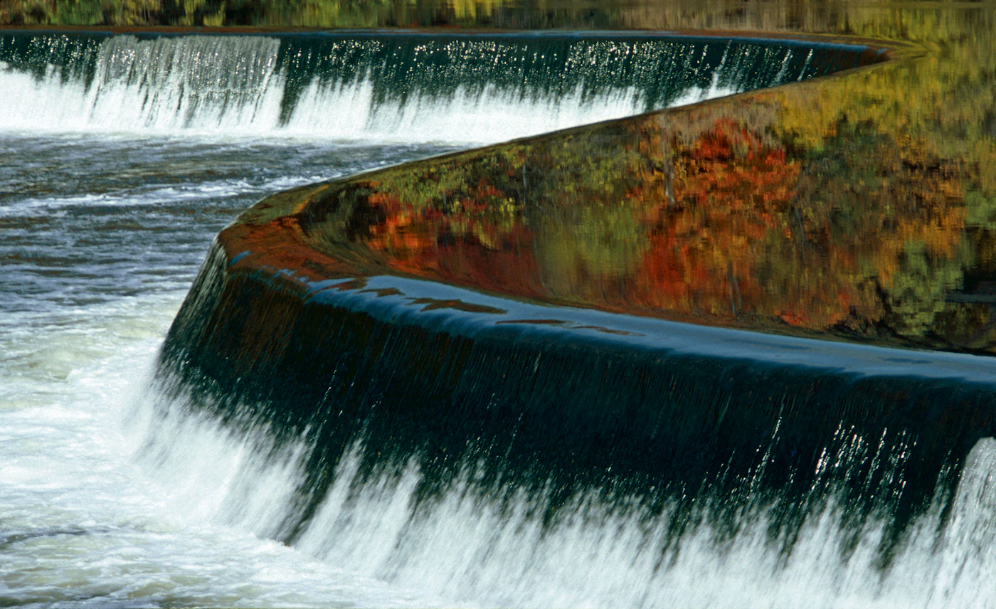 A wide waterfall cascading over a stone dam with fall foliage reflected in the churning water below in autumn