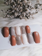 A collection of nail samples in various colors, including shades of brown, gray, beige, and a sparkling glitter finish, are arranged on a white surface. In the background, a cluster of dried flowers adds texture and contrast.