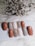 A collection of nail samples in various colors, including shades of brown, gray, beige, and a sparkling glitter finish, are arranged on a white surface. In the background, a cluster of dried flowers adds texture and contrast.
