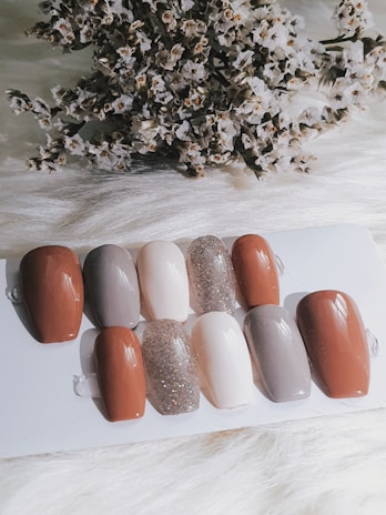 A collection of nail samples in various colors, including shades of brown, gray, beige, and a sparkling glitter finish, are arranged on a white surface. In the background, a cluster of dried flowers adds texture and contrast.