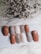 A collection of nail samples in various colors, including shades of brown, gray, beige, and a sparkling glitter finish, are arranged on a white surface. In the background, a cluster of dried flowers adds texture and contrast.