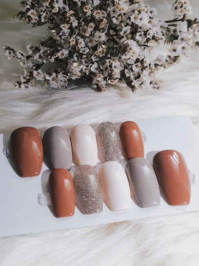 A collection of nail samples in various colors, including shades of brown, gray, beige, and a sparkling glitter finish, are arranged on a white surface. In the background, a cluster of dried flowers adds texture and contrast.