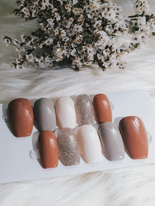 A collection of nail samples in various colors, including shades of brown, gray, beige, and a sparkling glitter finish, are arranged on a white surface. In the background, a cluster of dried flowers adds texture and contrast.