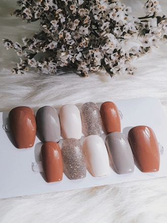A collection of nail samples in various colors, including shades of brown, gray, beige, and a sparkling glitter finish, are arranged on a white surface. In the background, a cluster of dried flowers adds texture and contrast.