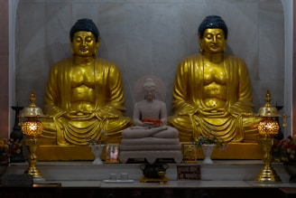 The image features two large golden Buddha statues seated in a meditative posture with a smaller white Buddha statue positioned in between them. The statues are adorned with intricate details and are placed on a platform alongside ornate lamps and decorative vases filled with flowers.