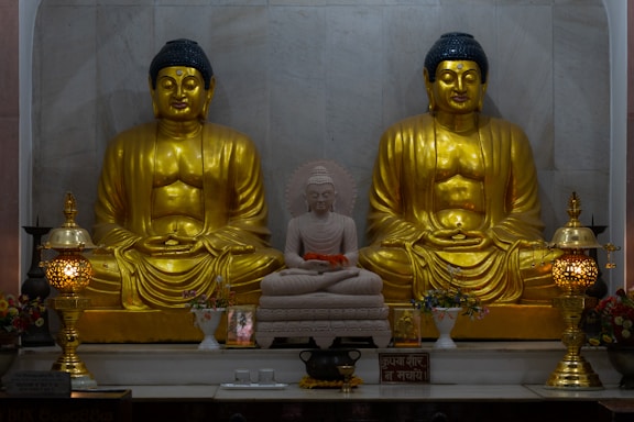 The image features two large golden Buddha statues seated in a meditative posture with a smaller white Buddha statue positioned in between them. The statues are adorned with intricate details and are placed on a platform alongside ornate lamps and decorative vases filled with flowers.