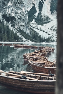 Wooden boats float peacefully on a clear lake surrounded by majestic, rocky mountains and lush evergreen trees. The scene reflects a serene and calm environment.