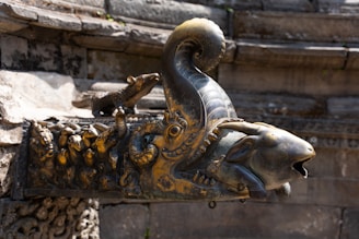 An intricately designed metal sculpture of a mythological creature adorns the corner of an ancient stone structure. The head resembles that of a goat with a curved horn, embellished with elaborate carvings and detailing. A small, stylized animal figure rests on its back, adding to the ornamental complexity. The overall appearance suggests a historical or cultural significance.