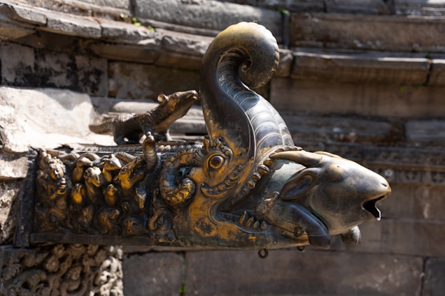 An intricately designed metal sculpture of a mythological creature adorns the corner of an ancient stone structure. The head resembles that of a goat with a curved horn, embellished with elaborate carvings and detailing. A small, stylized animal figure rests on its back, adding to the ornamental complexity. The overall appearance suggests a historical or cultural significance.