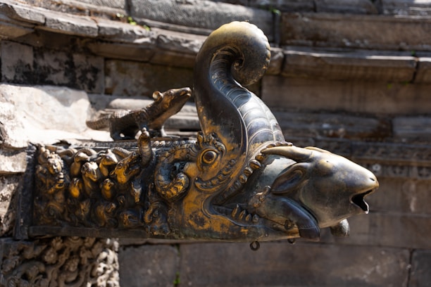 An intricately designed metal sculpture of a mythological creature adorns the corner of an ancient stone structure. The head resembles that of a goat with a curved horn, embellished with elaborate carvings and detailing. A small, stylized animal figure rests on its back, adding to the ornamental complexity. The overall appearance suggests a historical or cultural significance.