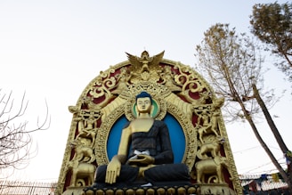 A large Buddha statue seated in a meditative pose is framed by ornate and intricately designed gold decorations with a blue background. There are also statues of elephants and mythical creatures incorporated into the frame. Tall trees are seen on either side of the statue.