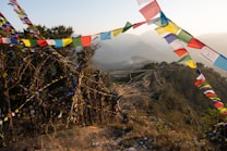 Brightly colored prayer flags are strung across the landscape, amidst a mountainous view with lush green hills. The flags flutter in a gentle breeze, creating a dynamic and spiritual ambiance. The background shows layers of distant mountains under a soft, hazy sky.