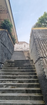 A staircase made of concrete and stones leads upwards. The stairway is bordered by concrete walls with a texture resembling a mesh pattern. On the left side at the top of the stairs, there is a house with a balcony featuring a lattice wooden railing and some potted plants. At the top of the stairs is a tiled rooftop with a geometric pattern. Trees with green foliage are visible on the right side, and the sky is clear and blue.
