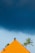 A vibrant orange-tiled roof sits beneath a dramatic dark sky, with a backdrop of tall palm trees swaying gently. The contrast between the bright roof and the ominous clouds creates a striking visual effect.