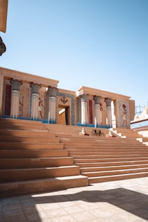 A grand stone staircase leads up to an Egyptian-inspired temple with intricate paintings and hieroglyphs covering the pillars and walls. Two identical statues flank the entrance, and a chair is positioned symmetrically between them. The architecture features traditional Egyptian motifs, including lotus-topped columns.