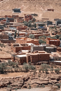 An arid landscape featuring a traditional village with red and brown buildings constructed from local materials. The village is spread across a rocky and dusty terrain, with sparse vegetation including a few trees and shrubs. Buildings vary in size and shape, creating a clustered layout that follows the natural contours of the land.