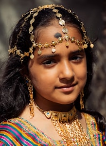 A young girl with long, dark hair adorned with elaborate gold jewelry. She is wearing a colorful, striped garment and a decorative headpiece made of gold and coins. The girl's expression is calm and serene, set against a blurred background.