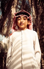 A young person wearing traditional clothing, including a white thobe and a red and white checkered keffiyeh. Palm trees are in the background, creating a natural setting.
