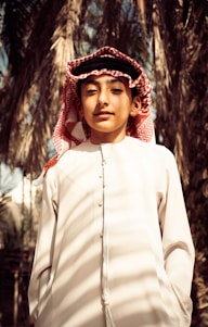 A young person wearing traditional clothing, including a white thobe and a red and white checkered keffiyeh. Palm trees are in the background, creating a natural setting.
