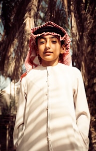 A young person wearing traditional clothing, including a white thobe and a red and white checkered keffiyeh. Palm trees are in the background, creating a natural setting.