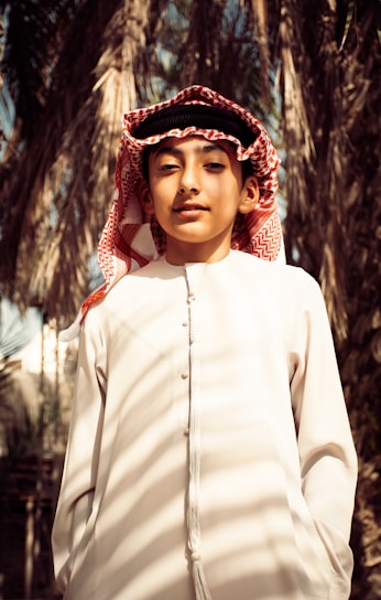 A young person wearing traditional clothing, including a white thobe and a red and white checkered keffiyeh. Palm trees are in the background, creating a natural setting.