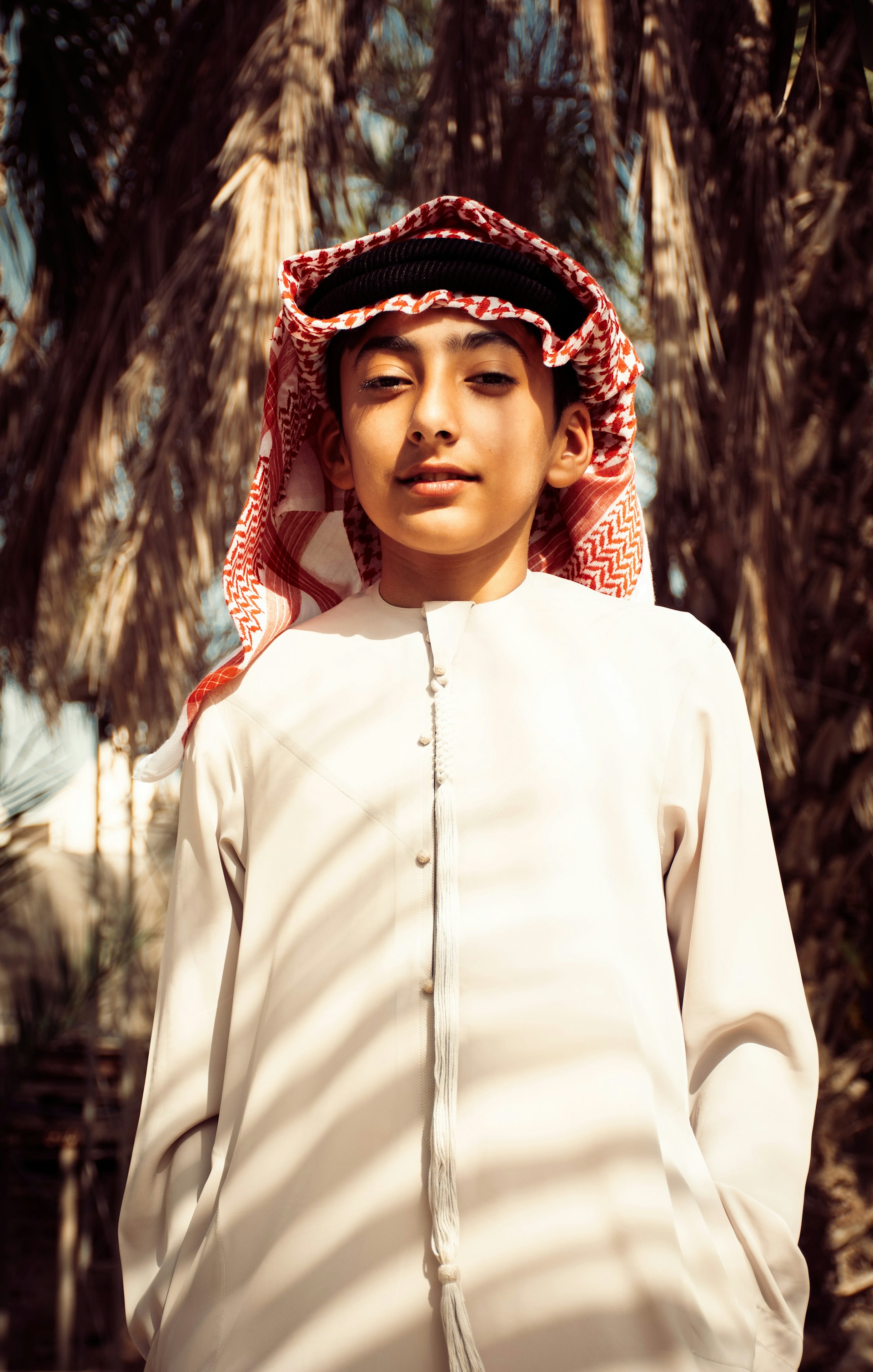 A young person wearing traditional clothing, including a white thobe and a red and white checkered keffiyeh. Palm trees are in the background, creating a natural setting.