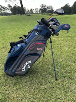 A blue and black golf bag with several golf clubs, including drivers and irons, is standing on a green grassy field. The brand name Callaway is prominently displayed on the side of the bag. Trees and bushes are visible in the distant background.