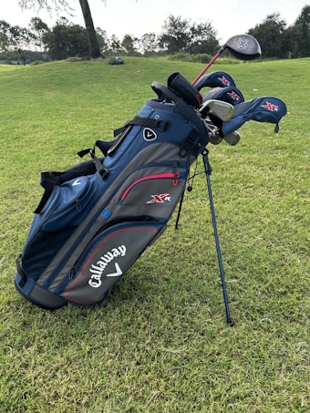 A blue and black golf bag with several golf clubs, including drivers and irons, is standing on a green grassy field. The brand name Callaway is prominently displayed on the side of the bag. Trees and bushes are visible in the distant background.
