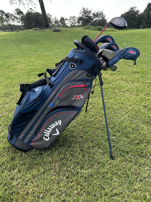 A blue and black golf bag with several golf clubs, including drivers and irons, is standing on a green grassy field. The brand name Callaway is prominently displayed on the side of the bag. Trees and bushes are visible in the distant background.