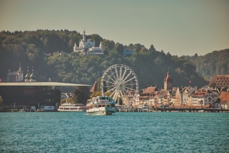 A scenic view of a lakeside town featuring a ferris wheel, historical buildings with ornate architecture, and forests in the background. A sightseeing boat sails in the foreground on the calm blue water, with hills and a large building atop them visible in the distance.