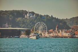 A scenic view of a lakeside town featuring a ferris wheel, historical buildings with ornate architecture, and forests in the background. A sightseeing boat sails in the foreground on the calm blue water, with hills and a large building atop them visible in the distance.
