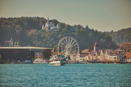 A scenic view of a lakeside town featuring a ferris wheel, historical buildings with ornate architecture, and forests in the background. A sightseeing boat sails in the foreground on the calm blue water, with hills and a large building atop them visible in the distance.