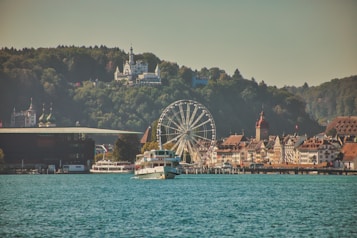 A scenic view of a lakeside town featuring a ferris wheel, historical buildings with ornate architecture, and forests in the background. A sightseeing boat sails in the foreground on the calm blue water, with hills and a large building atop them visible in the distance.