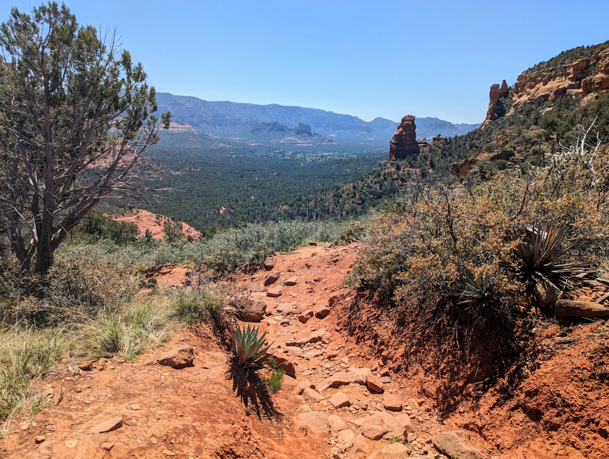 A red dirt trail through junipers and agave, looking down on a valley ringed by red rock spires
