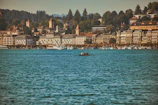 A large body of water with a team rowing a boat in the center. The background shows a cityscape with historic buildings, including hotels and homes with red-roofed towers, set against a backdrop of lush, green hills with dense tree cover. Numerous boats are visible along the shoreline.