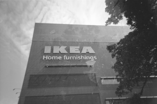 A large building facade with the word 'IKEA' prominently displayed above 'Home furnishings' in bold lettering. The image is in black and white, suggesting a timeless or vintage quality. Trees with dense foliage partially obscure the right side of the building, while wispy clouds fill the sky in the background.