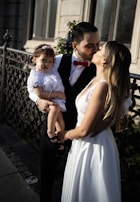 A couple is sharing a kiss while a man holds a young child dressed in white. They are standing by a decorative wrought iron fence, and the man is wearing a black suit with a red bow tie, while the woman is in a white bridal dress.