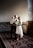A couple standing together in formal attire with the man holding a child. The woman is wearing a white dress and holding a bouquet of flowers, while the man is dressed in a suit with a red bow tie. The atmosphere is elegant with soft lighting and ornate architectural details in the background.