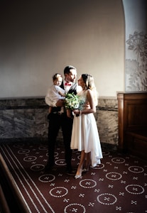 A couple standing together in formal attire with the man holding a child. The woman is wearing a white dress and holding a bouquet of flowers, while the man is dressed in a suit with a red bow tie. The atmosphere is elegant with soft lighting and ornate architectural details in the background.