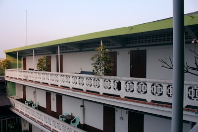 A multi-level building with a simple, utilitarian design. It features a green roof and white walls with symmetrical decorative patterns. The structure has balconies on each floor, lined with plants and small tables with chairs. Doors are evenly spaced along the walls, and there is minimalistic exterior decoration.