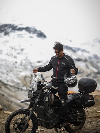A person in a black and red jacket stands next to a rugged motorcycle on a mountainous landscape with snow-covered peaks in the background. The individual holds a helmet and wears sunglasses, giving an adventurous impression.