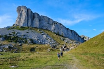 Two hikers are walking on a grassy path toward a village nestled at the base of a towering rocky cliff. A metal art structure is placed along the path. The weather is clear with blue skies, and the scene is surrounded by lush green hills and rocky terrain.