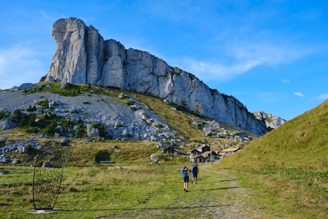 Two hikers are walking on a grassy path toward a village nestled at the base of a towering rocky cliff. A metal art structure is placed along the path. The weather is clear with blue skies, and the scene is surrounded by lush green hills and rocky terrain.