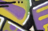 Bright and colorful graffiti featuring abstract shapes and bold black outlines, with a mix of purple, yellow, and green colors. The design includes dots and lines, presenting a vibrant and dynamic pattern.