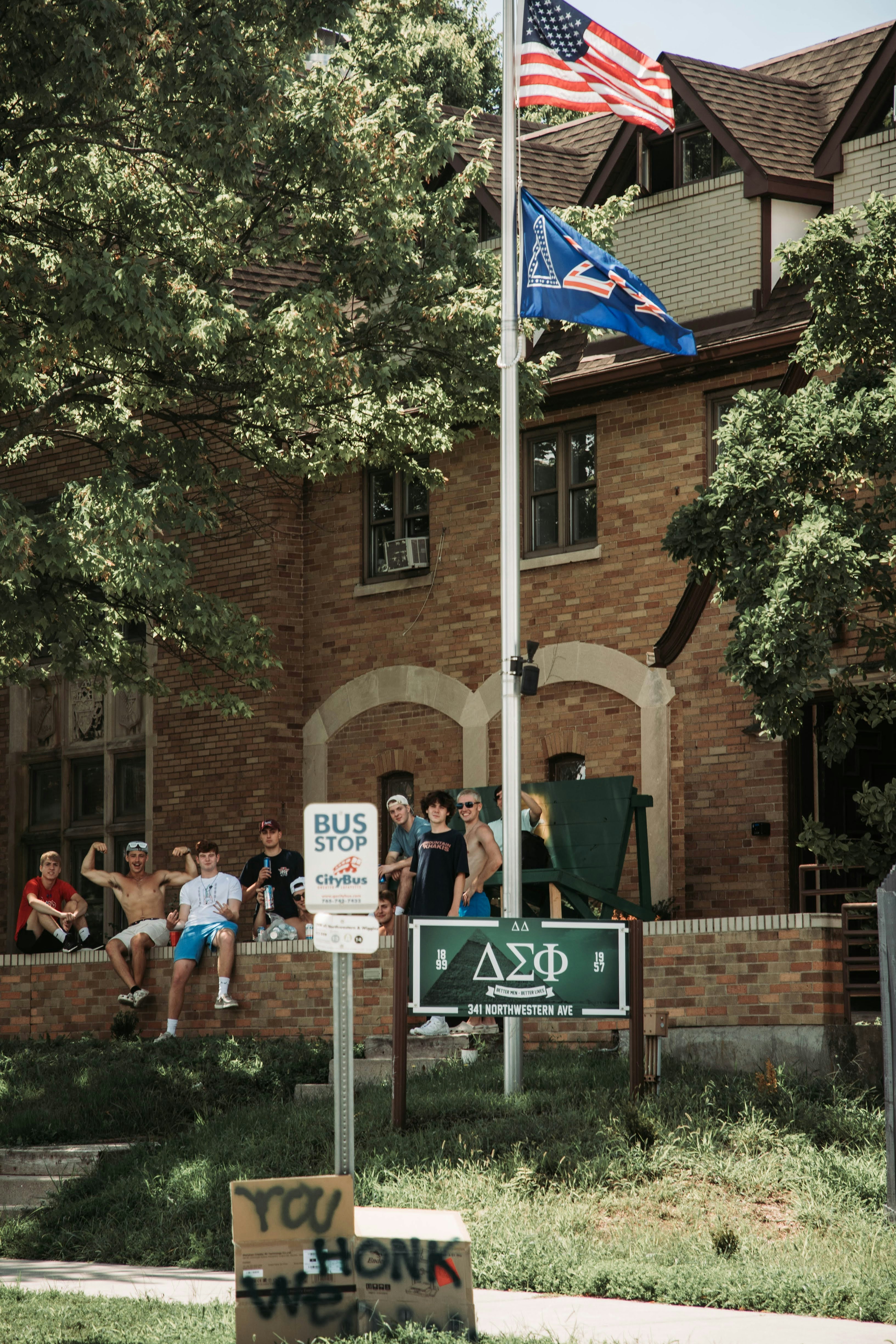 A group of young men are gathered on the lawn in front of a brick building, likely a fraternity house. Two flags, including an American flag, are flying atop a flagpole. A bus stop sign is visible on the sidewalk, along with a handmade sign that reads 'HONK WE'. Trees provide shade to the area.