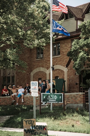 A group of young men are gathered on the lawn in front of a brick building, likely a fraternity house. Two flags, including an American flag, are flying atop a flagpole. A bus stop sign is visible on the sidewalk, along with a handmade sign that reads 'HONK WE'. Trees provide shade to the area.