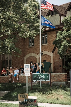 A group of young men are gathered on the lawn in front of a brick building, likely a fraternity house. Two flags, including an American flag, are flying atop a flagpole. A bus stop sign is visible on the sidewalk, along with a handmade sign that reads 'HONK WE'. Trees provide shade to the area.