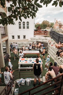 A large group of people are gathered around two tables in an outdoor courtyard. Many are standing while others are seated on stone steps, with trees and buildings in the background. The group appears to be socializing or playing games, and some are wearing sports jerseys. There are boxes of drinks placed on the ground.