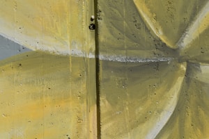 A textured concrete wall painted with a geometric design in shades of yellow and gray. The surface shows visible cracks and small holes, suggesting an aged or weathered appearance.