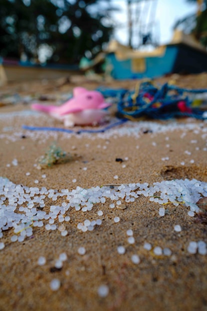 Small white plastic pellets scattered on a sandy beach, with blurred objects in the background including a pink figurine and assorted debris.